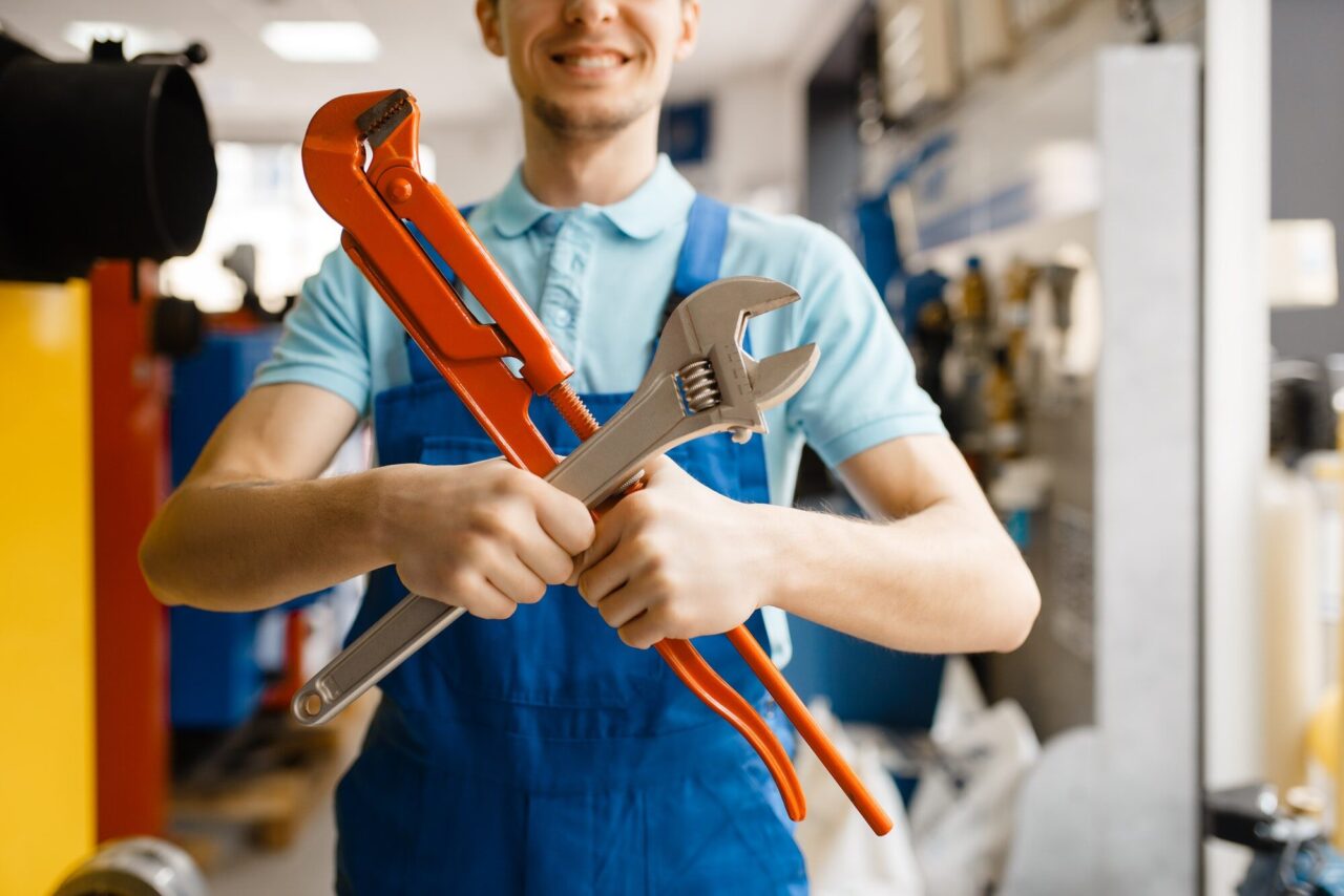 A man in work overalls smiles while holding a red pipe wrench and an adjustable wrench in a workshop setting. Tools and equipment are visible in the background.