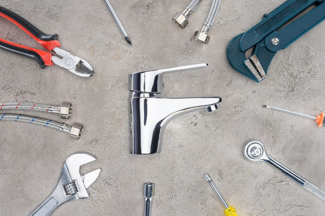 A shiny chrome faucet is surrounded by various plumbing tools and accessories laid out on a concrete surface. The arrangement highlights the tools commonly used for plumbing tasks.
