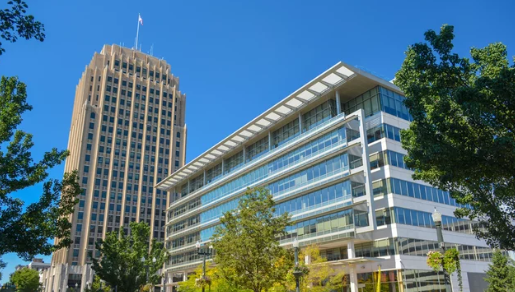 Two distinct buildings under a clear blue sky a tall, classic skyscraper on the left and a modern, glass-fronted structure on the right, surrounded by greenery